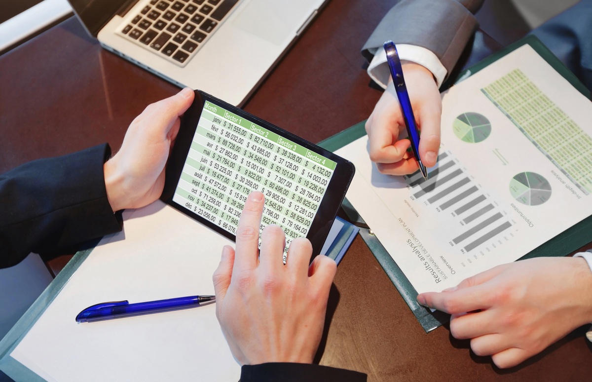 Manos sobre una tablet que muestra una hoja de cálculo de Excel, junto a informes con gráficos
